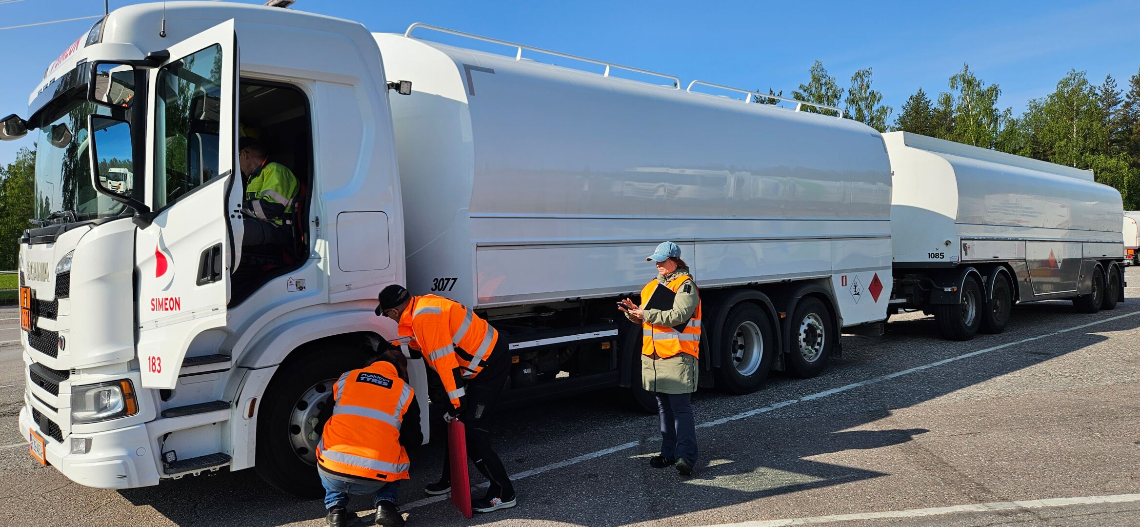 En hvit Simeon-lastebil med to tankvogner står parkert, mens tre Vianor-arbeidere i oransje sikkerhetsvester inspiserer dekkene.