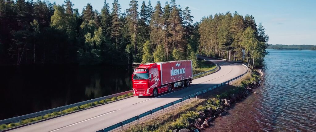 Henkilö huomiovärisessä oranssissa takissa ja mustassa pipossa seisoo punaisen kuorma-auton vieressä lumisessa ulkoympäristössä.