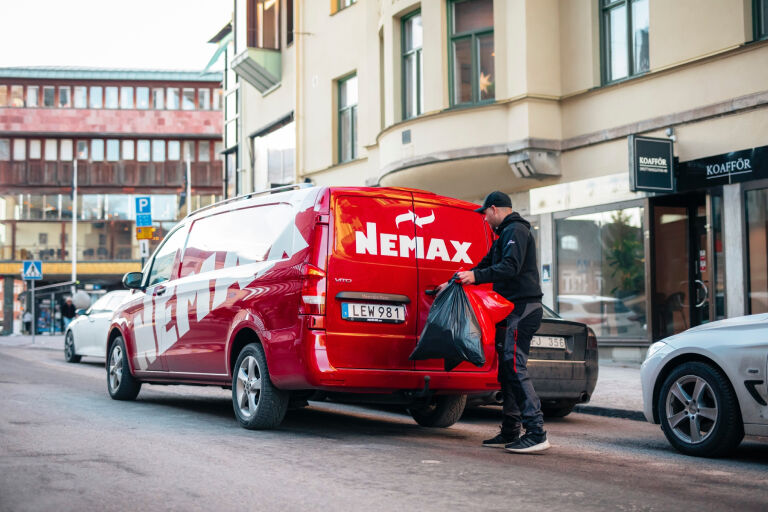 Red Nemax service van parked on a city street, with a person loading a large black garbage bag into the back.