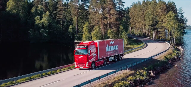 Röd transportbil kör längs en böjd sjöväg kantad av skog, med vatten till höger och täta träd i bakgrunden.