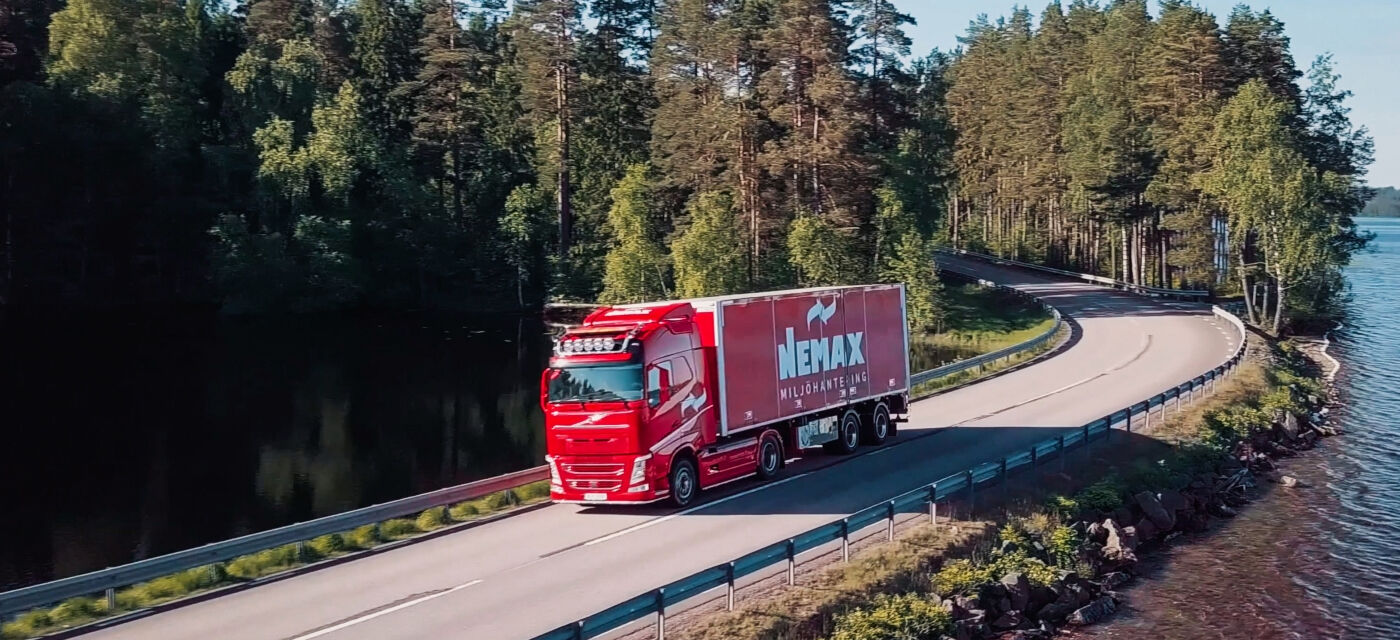 Röd transportbil kör längs en böjd sjöväg kantad av skog, med vatten till höger och täta träd i bakgrunden.