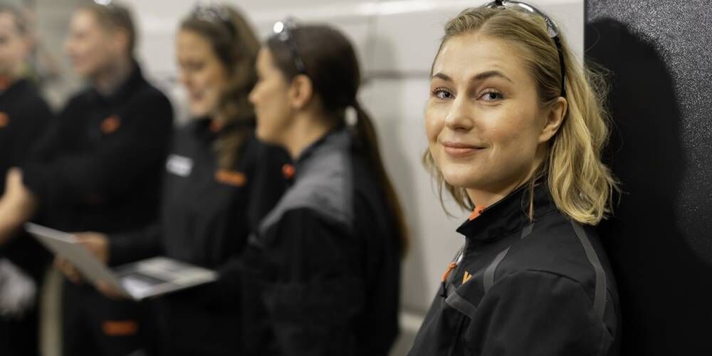 Kvinnelig mekaniker smiler mot kamera foran teammøte hos Vianor verksted.