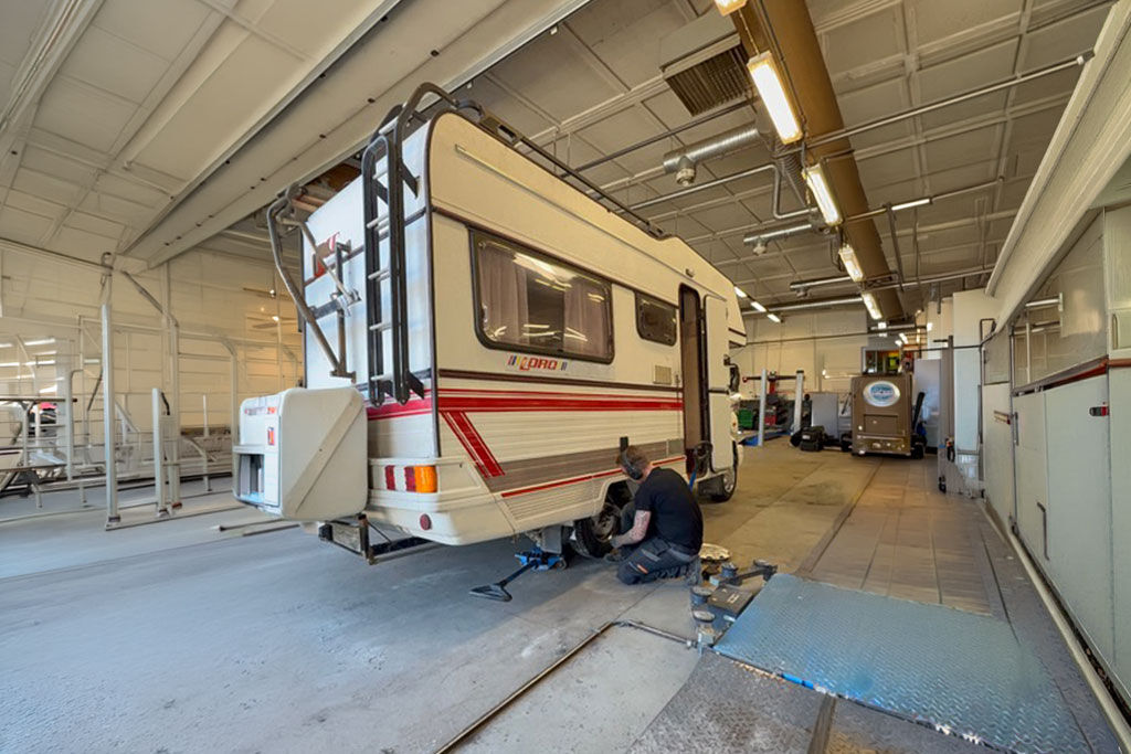 Asuntoauto on pysäköitynä korjaamohalliin, ja asentaja on polvistuneena takapyörän vieressä vaihtamassa rengasta. Työkaluja ja tunkki on asetettu lattialle lähelle.  