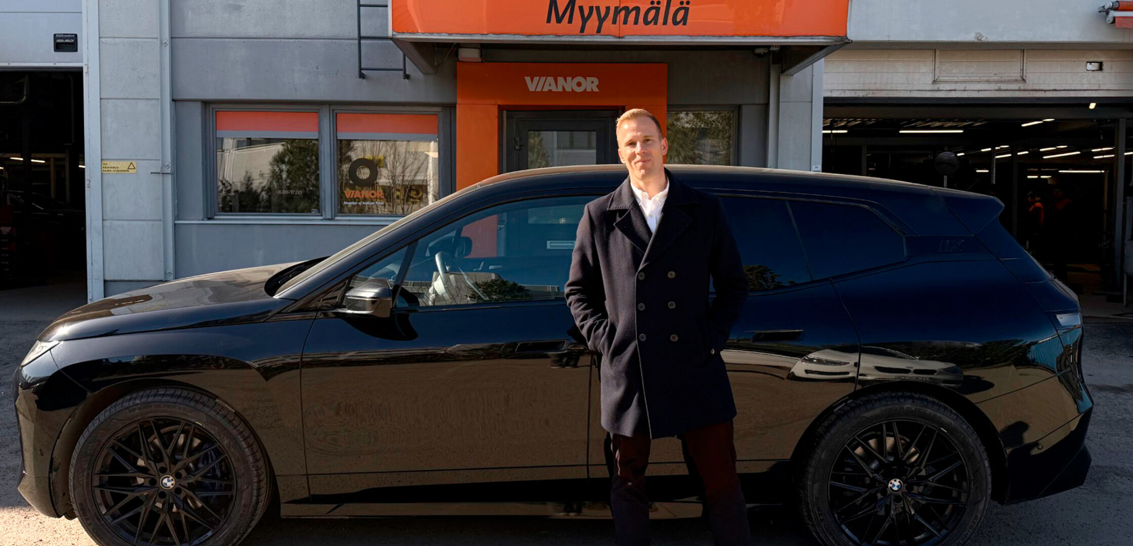 Musta BMW iX ‑sähkö-SUV pysäköitynä rannalle; aurinkolaseja käyttävä henkilö seisoo auton vieressä aurinkoisena alkukevään päivänä. 