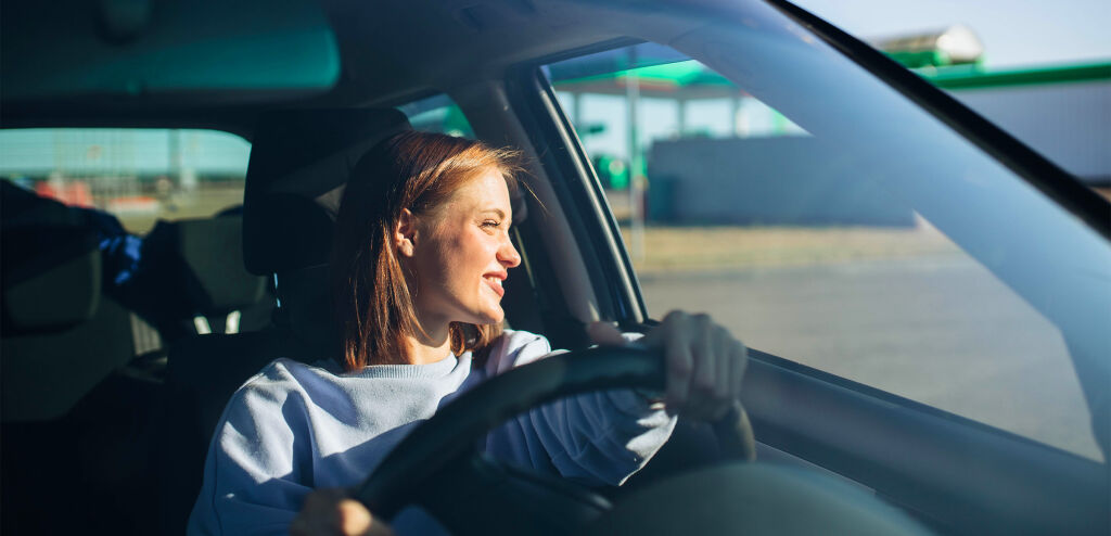 Driver inside a car behind the steering wheel smiling and looking out of the car window with sun shining on her face