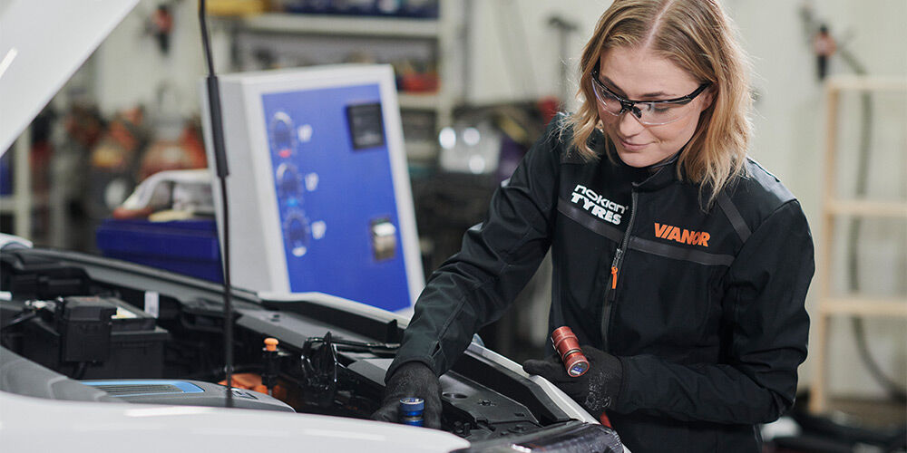 En kvinnelig Vianor-tekniker inspiserer motorrommet på en elbil og holder en lykt i hånden. Hun har på seg vernebriller og arbeidstøy med Nokian Tyres og Vianor-logoer.