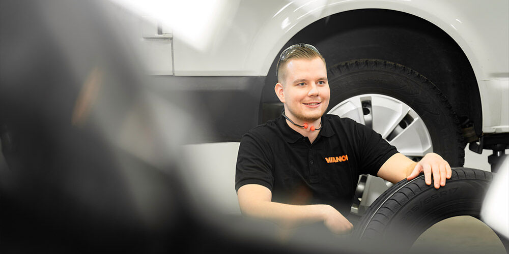 Hymyilevä mekaanikko mustassa Vianor-paidassa pitelee sähköauton rengasta korjaamolla. Taustalla näkyy valkoinen auto ja toinen asennettu rengas.