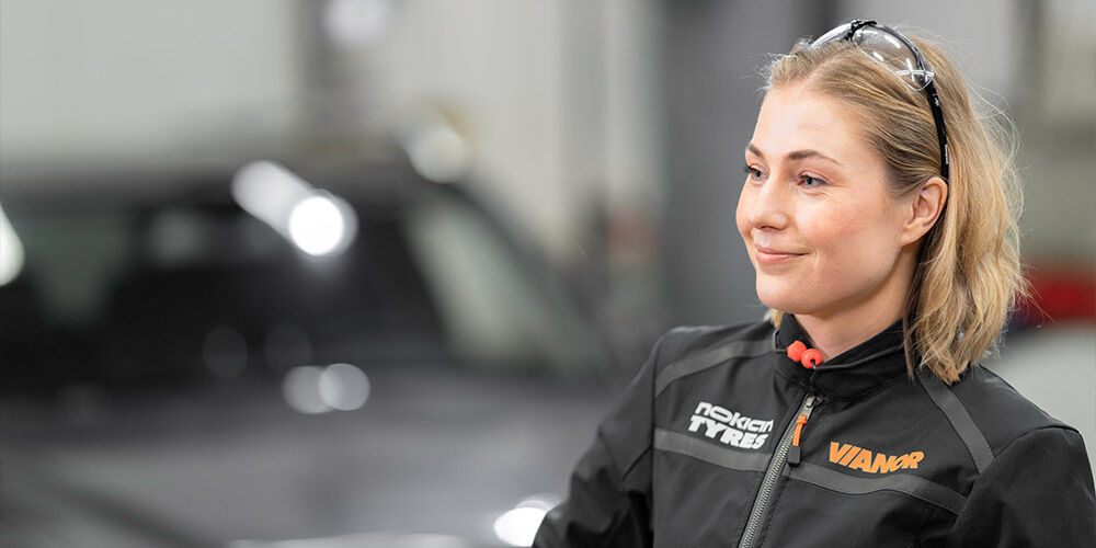 Hymyilevä Vianor-työntekijä mustassa Nokian Tyres -takissa seisoo korjaamolla. Hänellä on suojalasit nostettuna hiuksiinsa ja auto näkyy taustalla.
