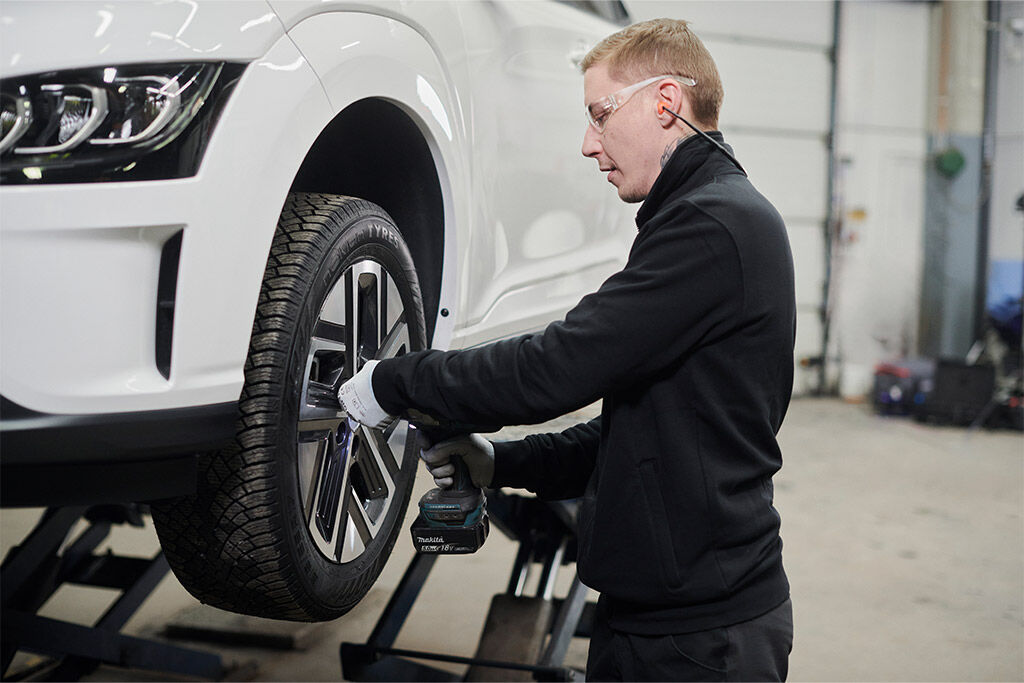 Mekaanikko asentaa sähköauton talvirengasta korjaamolla. Valkoinen sähköauto on nosturilla, ja renkaan kuviointi näkyy selvästi.