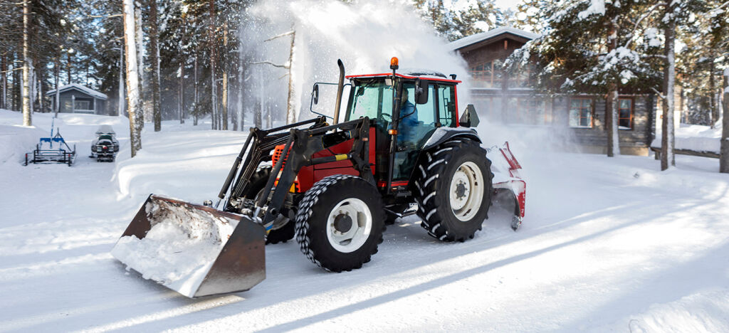 En rød traktor med frontlaster og snøfreser festet bakpå rydder snø i et skogkledd område