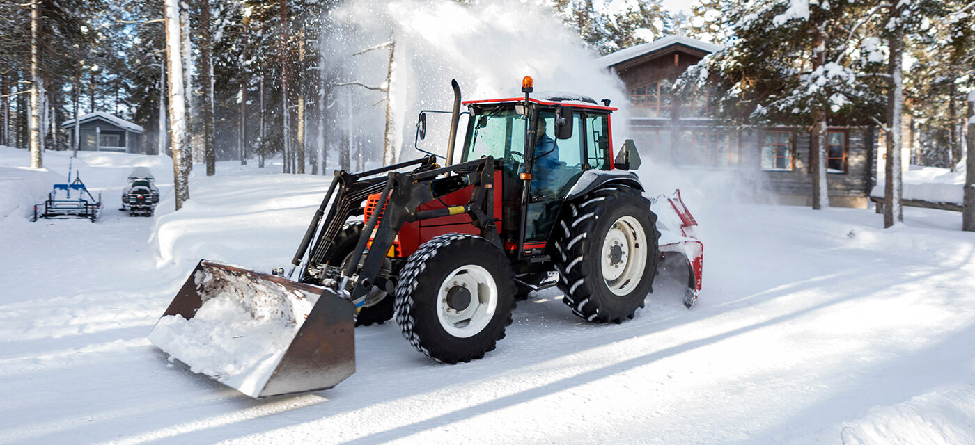 En rød traktor med frontlaster og snøfreser festet bakpå rydder snø i et skogkledd område