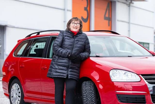 Nainen nojaamassa punaisen auton konepeltiin rakennuksen pihassa