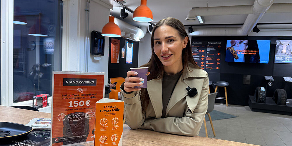 Person wearing a light trench coat and dark outfit leaning on the service desk at Vianor service center with coffee cup in hand