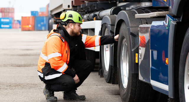 En person i verneklær og med vernehjelm inspiserer dekkene på en lastebil med containere i bakgrunnen.